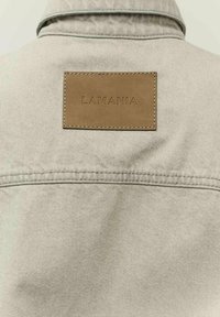 Beige farmerdzseki, amelyen egy barna bőr folt található, rajta a „LAMANIA” felirat domborítva, a hátsó felső részen. A gallér és a varratok láthatóan meg vannak varrva.