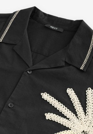 Black shirt with a collar, featuring white embroidered floral design, two black buttons, and contrasting stitching along the collar.