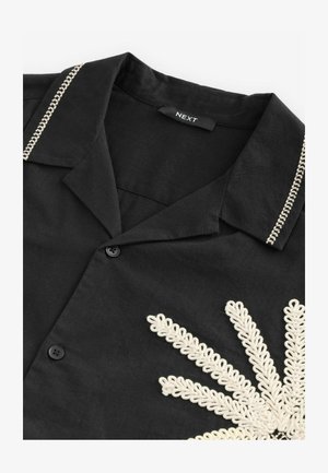 Černá košile s límcem, zdobená bílou výšivkou květinového vzoru, dvěma černými knoflíky a kontrastním prošíváním kolem límce.