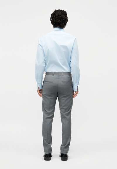 Άνδρας με σγουρά σκούρα μαλλιά που φοράει ανοιχτό μπλε πουκάμισο και γκρι παντελόνι, στέκεται γυρισμένος πλάτη, σε απλό λευκό φόντο.