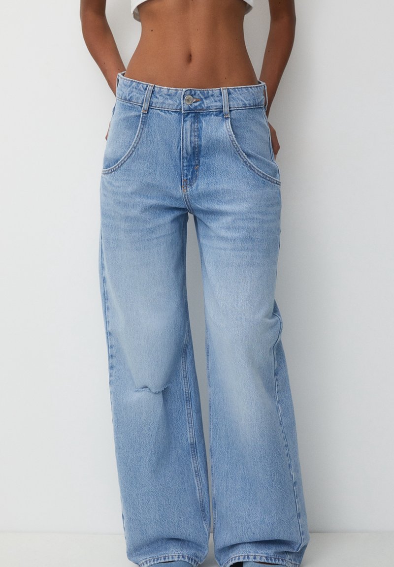 Femme portant un jean bleu clair taille haute à jambes larges avec des poches avant et une petite déchirure sur la cuisse gauche, debout devant un fond blanc uni.