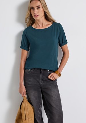 Teal kortærmet top med optrullede manchetter, parret med mørkegrå jeans. Bemærkelsesværdig glat tekstur; model der holder en tan ruskind taske.