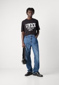 Black T-shirt with white "Hugo Creative Dept." graphic, blue denim jeans, black shoes, and a black crossbody bag. Casual fit and style.