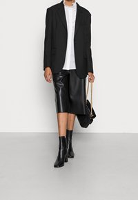 Blazer nero su una camicia bianca a balze, abbinato a culotte in pelle nera, stivali neri alla caviglia e una borsa nera con tracolla a catena dorata.