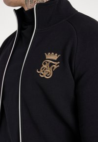 Giacca nera con zip e colletto alto. Presenta un logo ricamato in oro e una corona sul petto. Realizzata in materiale morbido ed elastico.