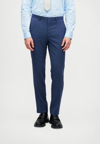 Άνδρας που φοράει καλά ραμμένα ναυτικά μπλε κοστούμια παντελόνια, ανοιχτό γαλάζιο πουκάμισο, μπεζ γραβάτα και μαύρα κομψά υποδήματα σε απλή φόντο.