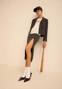 Blazer gris ajusté avec poches avant, associé à une jupe grise à fente haute. Le modèle porte un t-shirt graphique blanc, des chaussettes blanches et des talons noirs.