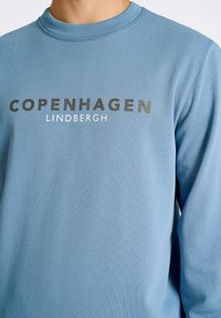 Blå sweatshirt av mjukt tyg med rund halsringning, ribbade ärmslut och djärv text "COPENHAGEN" och "LINDBERGH" i kontrasterande färger.