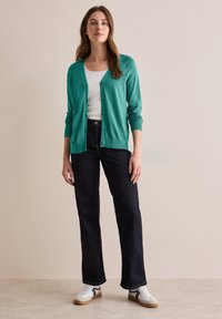 Teal cardigan met knopen, gedragen over een witte tanktop. Gecombineerd met donkere spijkerbroek en witte sneakers met zwarte accenten.