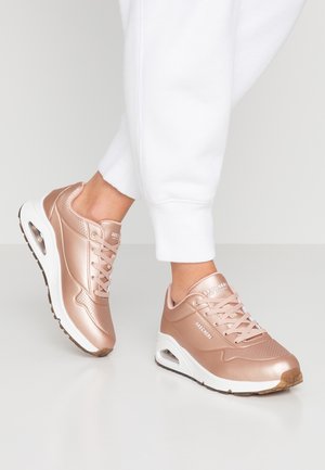 Persoon die rosegouden Skechers-sneakers draagt met witte zolen en luchtkussenhielen, gecombineerd met witte broeken tegen een lichte achtergrond.