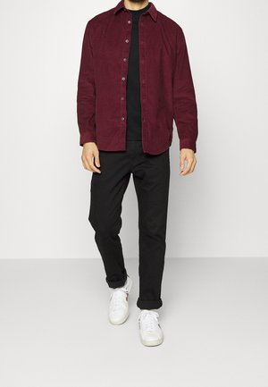 Burgundy corduroy-paita napilla edessä, musta t-paita alla, yhdistetty mustiin housuihin ja valkoisiin tennareihin. Sujuva rakenne, rento istuvuus.