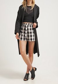 Nainen, jolla on päällään musta verkkop crop-top, pitkä musta takki, mustavalkoiset ruutushortsit ja mustat avokärkiset nilkkabootsit, seisoo jalat ristissä.