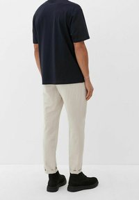 Schwarzes T-Shirt mit kurzen Ärmeln, cremefarbene, taillierte Hose und schwarze Knöchelstiefel, von hinten vor einem weißen Hintergrund betrachtet.