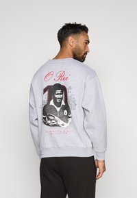 Sudadera gris con un gráfico en la parte posterior que muestra a un jugador sosteniendo un balón de fútbol, el texto "O Rei" y el número "10".