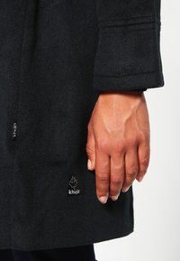 Cappotto di lana nero con tessuto texturizzato, caratterizzato da un'etichetta logo visibile e dettagli di cucitura evidenti sulla manica. Mano appoggiata sul tessuto.