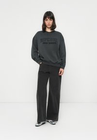Donna con i capelli scuri e lisci, che indossa un sweatshirt nera "Ripcurl Surf Sports", pantaloni larghi neri e scarpe piatte argentate, in piedi contro un muro bianco.