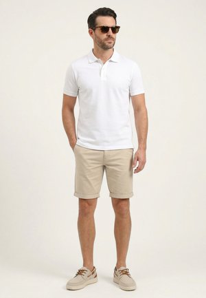 Mann mit Sonnenbrille, weißem Poloshirt, beigen hochgekrempelten Shorts und beigen Freizeitschuhen, der mit einer Hand in der Tasche steht.