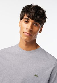 Grå bomullströja med rundad hals och en liten grön broderad alligatorlogga på vänster bröst. Mjuk textur, enkel design.