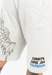 Heledad hallika lühikesed püksid, millel on suur tiiva graafika vasakul küljel ning tekst "PIIRID ON ILLUSIOON" julgete mustade ja siniste fontidega.