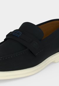Μαύρο παπούτσι loafer τύπου slip-on με λευκή σόλα, ραμμένες λεπτομέρειες και διακοσμητικό λουράκι με μικρή μπλε αγκράφα στην κορυφή.