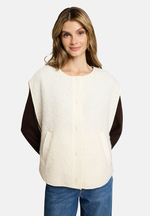 Giovane donna che indossa un gilet bianco senza maniche con bottoni sopra una camicia scura a maniche lunghe e jeans blu, in piedi davanti a uno sfondo bianco.
