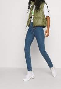 Žena v modrých skinny džínech, bílém tričku s grafickým potiskem, olivově zelené péřové vestě a bílých teniskách, která kráčí proti neutrálnímu pozadí.