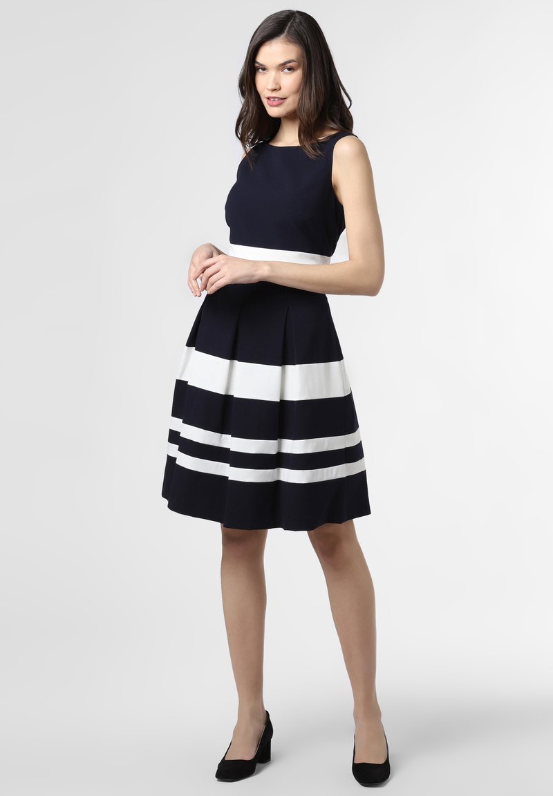 Ein marineblaues Kleid mit tailliertem Oberteil und ausgestelltem Rock, das horizontale weiße Streifen aufweist und aus einem glatten Stoff gefertigt ist. Kombiniert mit schwarzen High Heels.