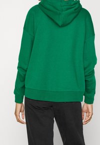 Sweat à capuche vert avec une poche kangourou, des poignets et un ourlet côtelés. Fabriqué en tissu doux avec une coupe décontractée et carrée.