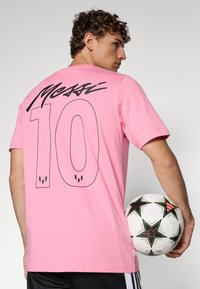 Pink cotton t-shirt with black "Messi" script and "10" on the back. Model is holding a soccer ball with a star pattern, wearing black shorts.