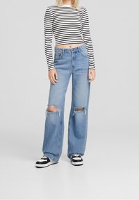 Donna che indossa jeans strappati a vita alta di colore blu chiaro, un crop top a strisce bianco e nere a maniche lunghe e sneakers bianche e nere, in piedi contro uno sfondo grigio.