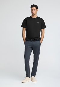 Schwarz Baumwoll-T-Shirt mit kurzen Ärmeln und einem kleinen Logo-Patch, kombiniert mit grauen Hosen und hellen Sneakers. Einfaches, lässiges Design.