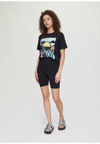 T-shirt noir avec un graphisme coloré représentant un soleil et des palmiers, associé à un short noir et des sandales bleues avec des fermetures à double boucle.