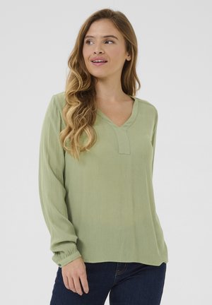 Blouse vert clair à manches longues avec un décolleté en V, tissu texturé et détails subtils aux poignets. Associée à un denim foncé.
