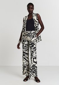 Schwarz-weiß gestreiftes ärmelloses Oberteil mit Zebra-Muster und weite Hose aus leichtem Stoff, kombiniert mit einem schwarzen Oberteil und Slippern.