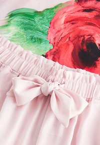 Lys pink nederdel med en rummelig talje og en bundet sløjfedetalje. Baggrunden prydes af abstrakte røde og grønne blomster mønstre.