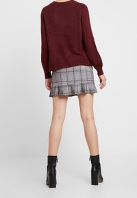 Pull en tricot bordeaux à manches longues, associé à une jupe grise à carreaux avec des volants. Bottines noires à talon épais.
