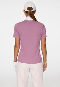 Kurzärmliges Polo-Shirt in hellem Lila, aus glattem Stoff, mit klassischem Schnitt. Der Kragen ist weiß, dazu werden helle Hosen und ein Golfhandschuh getragen.