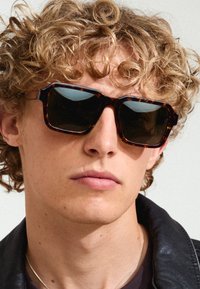 Lunettes de soleil en écaille de tortue avec des verres foncés, de forme rectangulaire et avec des montures épaisses. Portées par une personne aux cheveux bouclés et portant une veste noire.