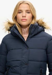 Veste matelassée bleu marine avec capuche en fausse fourrure amovible, design matelassé et fermeture éclair à l'avant. Dotée de poches latérales et de poignets élastiques.