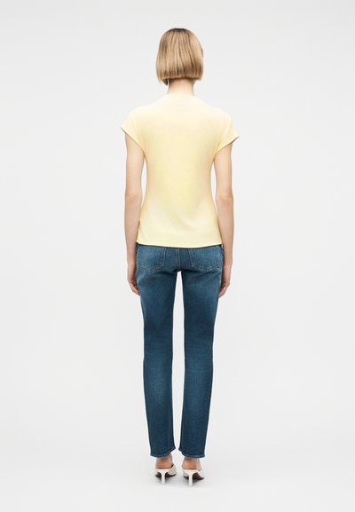 Γυναίκα με κοντά ξανθά μαλλιά, που φοράει ανοιχτόκίτρινη μπλούζα με κοντά μανίκια, μπλε τζιν και λευκές ψηλοτάκουνες γόβες, που στέκεται γυρισμένη προς τα πίσω.