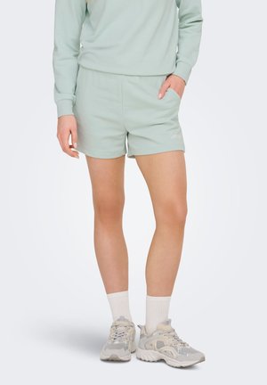 Vrouw draagt een lichtgroene sweatshirt en bijpassende korte broek met witte sokken en lichtgrijze sneakers, staat met één hand in de zak.