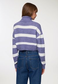 Kobieta widziana od tyłu, ubrana w fioletowy i biały sweter z golfem w paski oraz dżinsy z wysokim stanem na jednolitym tle.