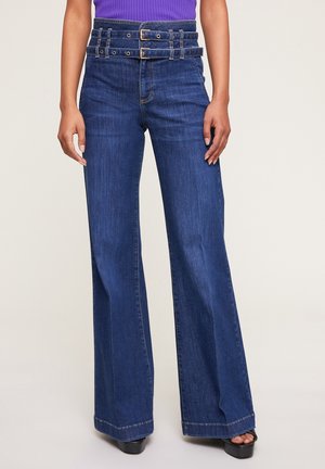 Kvinde iført højtaljede, viddeben, mørkeblå jeans med dobbelt bælte-detalje, kombineret med en lilla ærmeløs top og sorte platformsandaler.