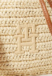 Sirkkakankaasta kudottu beige laukku, jossa on teksturoitu pinta. Sillä on silmiinpistävä logo kudottuna materiaaliin ja ruskeat nahkakahvat.