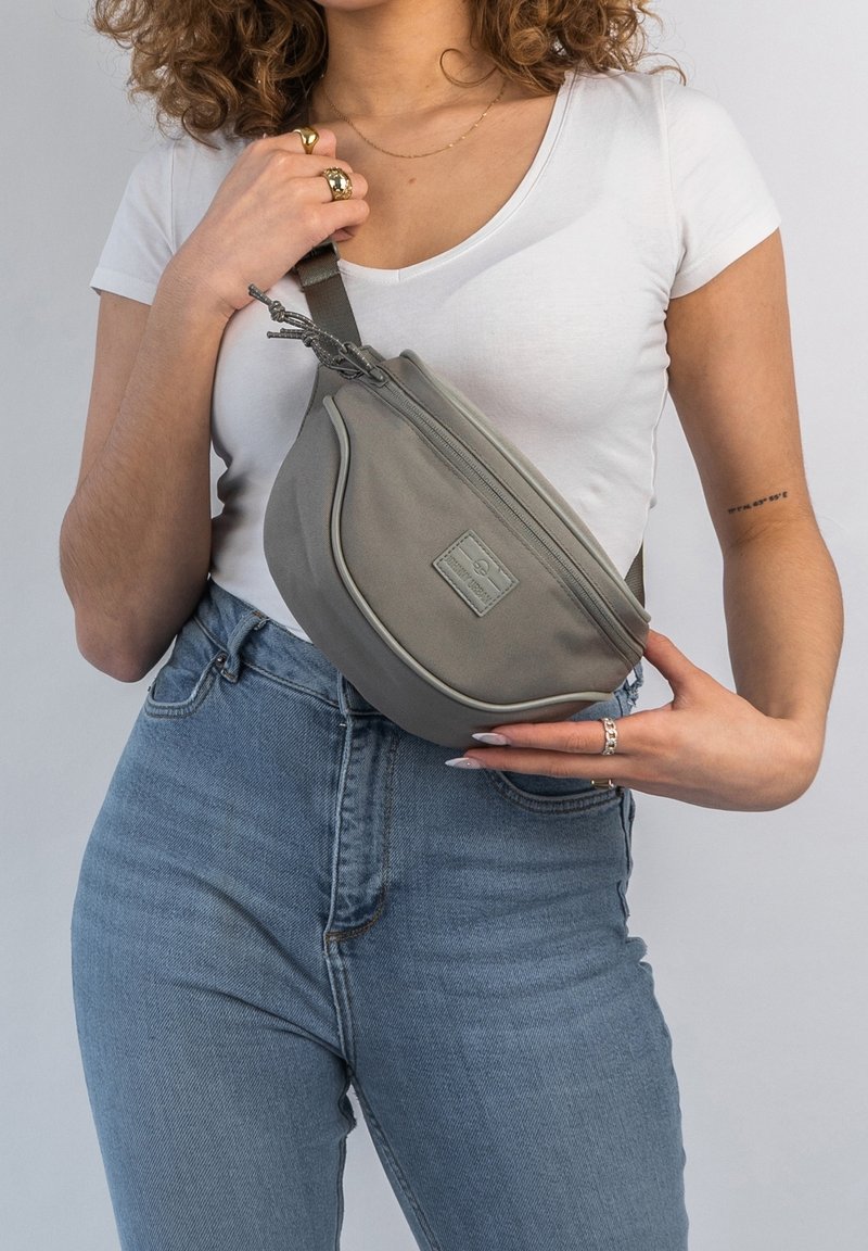 Femme portant un t-shirt blanc et un jean tient un sac bandoulière gris avec fermeture éclair sur sa poitrine, sur fond clair.