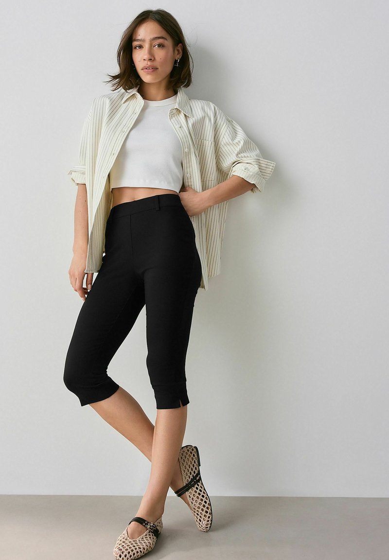 Svarta cropped byxor med smal passform, matchade med en vit crop top och en ljusrandig skjorta med knappar; strukturerade, mönstrade skor fulländar looken.