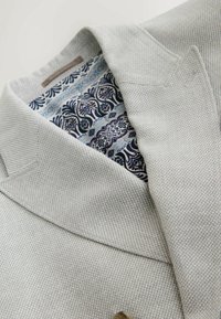 Veste blazer gris clair texturée avec une doublure intérieure fleurie bleue et noire à motifs, et une étiquette marron à l'intérieur du col.
