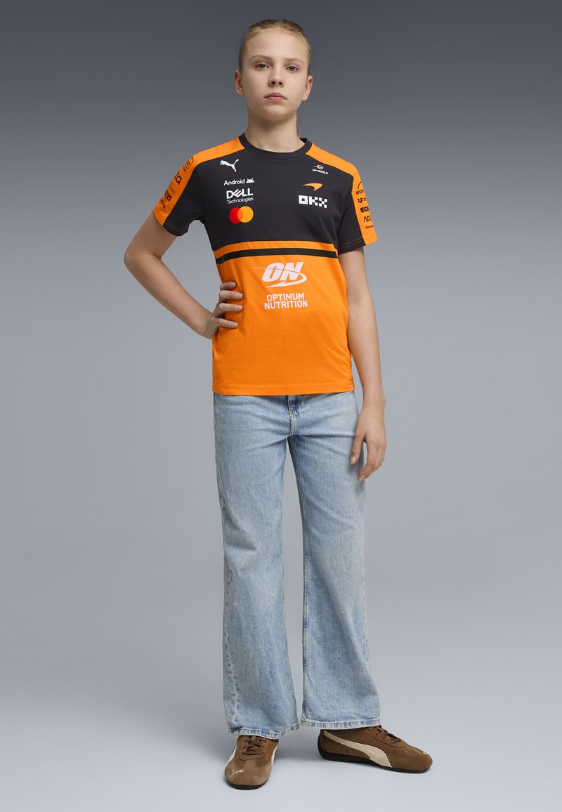 Jeune personne debout, main sur la hanche, portant un maillot de sport noir et orange avec des logos de sponsors, un jean bleu et des baskets marron.