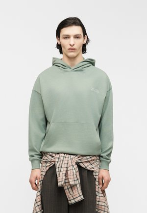 Ung person med mørkt hår iført salviegrøn hættetrøje og ternet skjorte bundet om livet, står foran en ensartet lys baggrund.