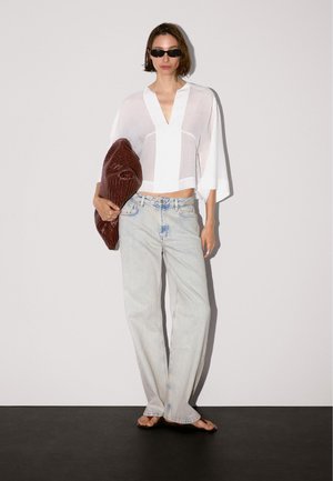 Camicia bianca, corta, trasparente con scollatura a V; jeans azzurri a gamba larga; borsa intrecciata marrone; occhiali da sole neri; sfondo minimale.